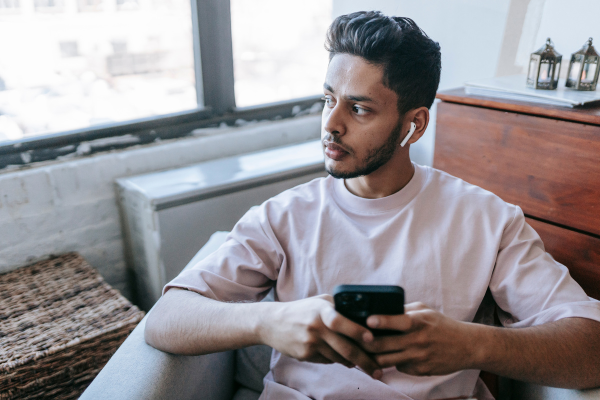 Thoughtful Indian man with earbuds browsing smartphone